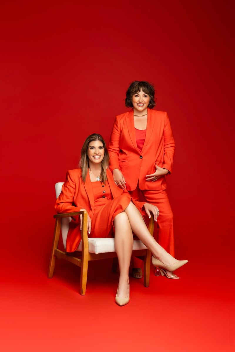 Maggie seated in an orange suit with Rachel standing behind her, against a red backdrop
