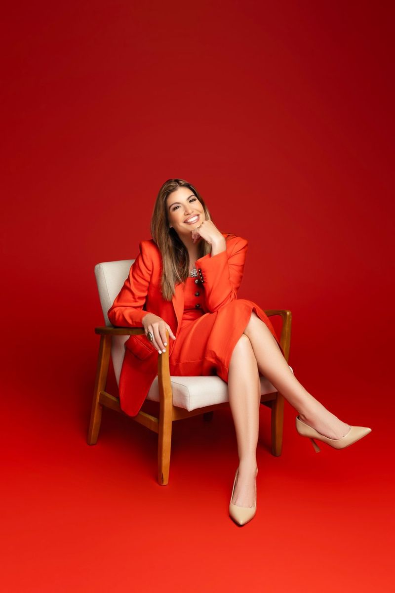 Maggie B. Hopkins, broker-owner, smiling in an orange suit seated in a white chair against a red backdrop